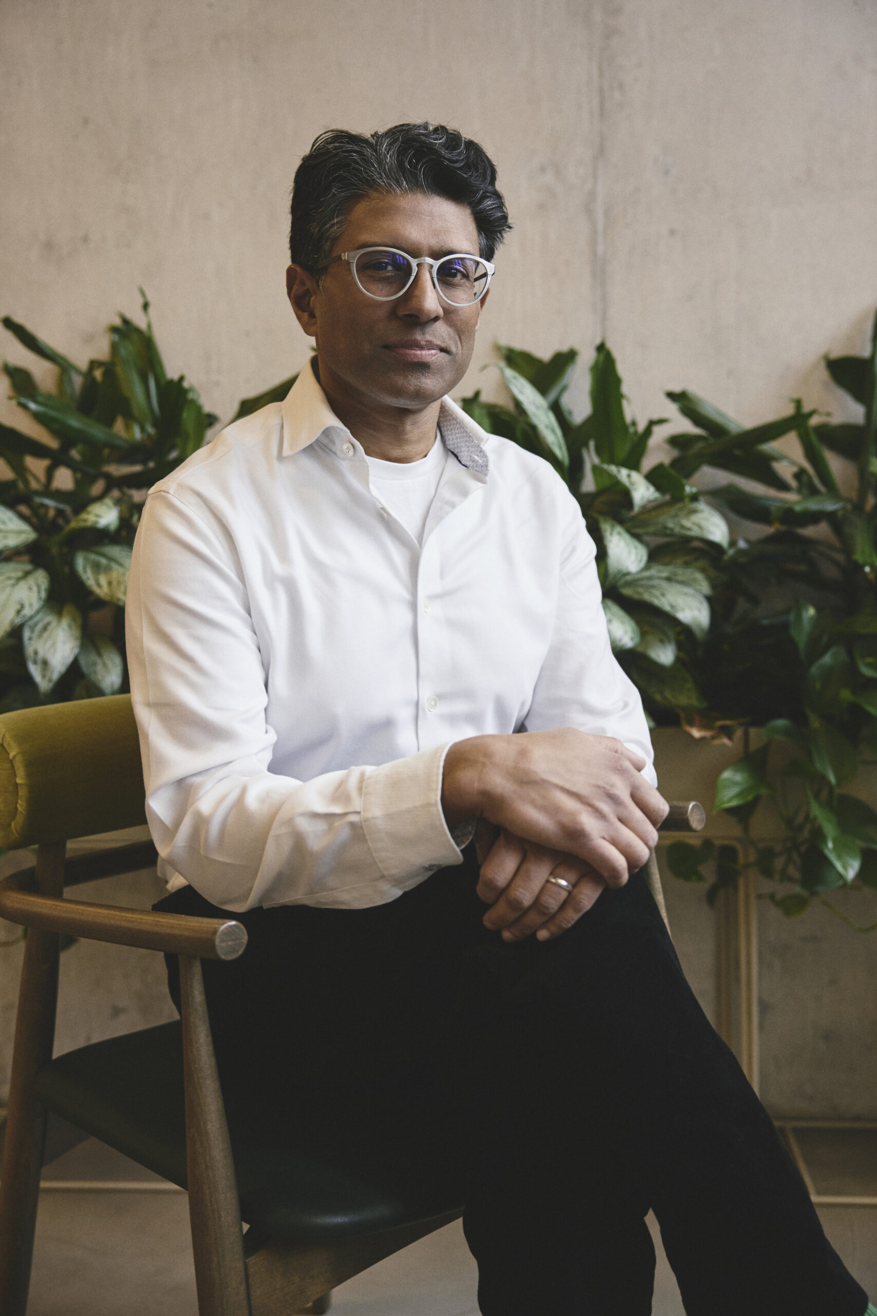 Khuzema Hussain sits in a modernist chair . There are indoor plants in the background and a concrete wall.