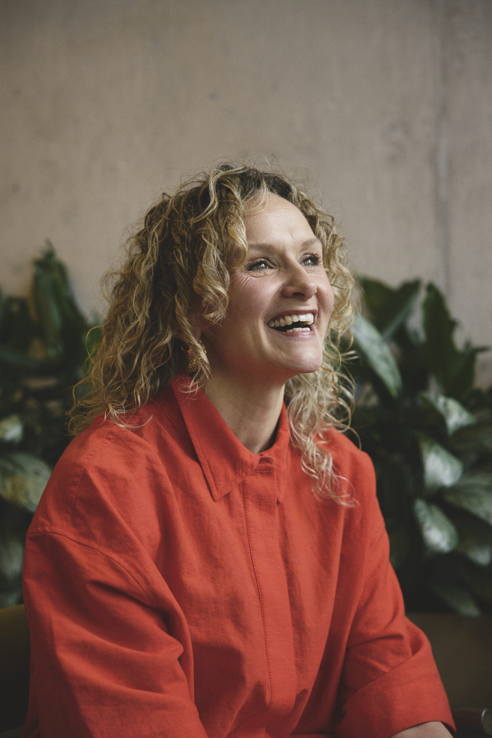 A blonde woman in a red shirt laughs. She is in a creative office space, with plants and a concrete wall.