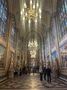 The interior of a tall gothic building. There are paintings on the wall and light comes in via tall windows.