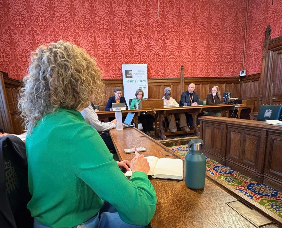A woman in green sits in the foreground of a traditional panelled room. A row of people sit along one side of a table in the background