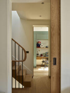 Looking across a top lit stairwell through tall doorways with pocket doors. In the far room is a desk and neat joinery cupboards.